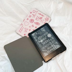 May include: A black Kindle e-reader with a gray case. The e-reader is open to a book titled "Before We Were Innocent" by Ella Berman. The case has a pink and white design with hearts, stars, and other small illustrations.