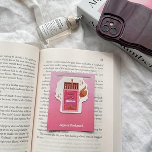 May include: A pink and white magnetic bookmark with a cartoon illustration of a matchbox with a flame. The matchbox has the words "Slow Burn Book Club" printed on it. The bookmark has the words "Magnetic Bookmark" printed on it.