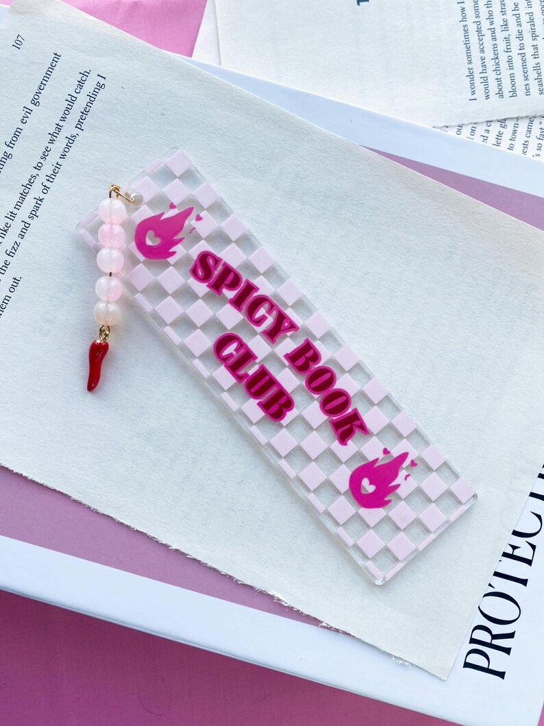 May include: Pink and white chequered bookmark with the text "Spicy Book Club" in pink lettering. A pink flame with a heart is on either end of the bookmark.