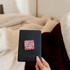 May include: A black e-reader with a pink and red "Smutty Book Club" sticker featuring strawberries and a heart design. The e-reader is held against a backdrop of a white and burgundy blanket.
