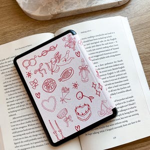 May include: A white e-reader with a black border, displaying a page of a book with red illustrations. The book is open and the e-reader is lying on top of it. The text on the page reads "Making colour" and "y knew, reading a storybook out loud to a should have been tucked up in bed."