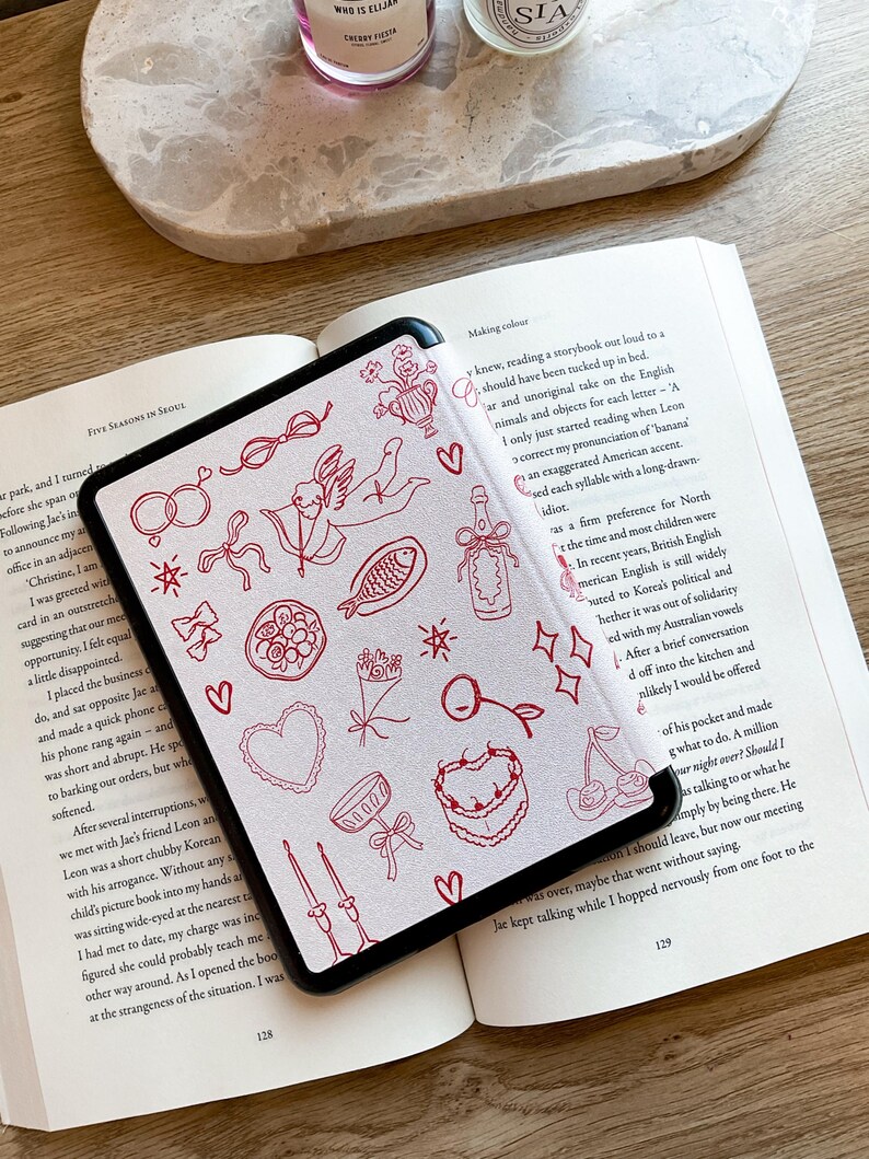 May include: A white e-reader with a black border, displaying a page of a book with red illustrations. The book is open and the e-reader is lying on top of it. The text on the page reads "Making colour" and "y knew, reading a storybook out loud to a should have been tucked up in bed."
