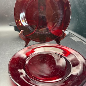 May include: Two round plates made of translucent deep red glass. One plate is positioned upright on a small wooden stand, while the second plate lies flat in the foreground. Both plates feature a smooth, reflective surface and a simple circular design.