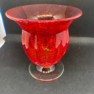 May include: A red glass vase with a textured, bubbled surface and a flared rim. The vase has a round base and a clear glass pedestal.