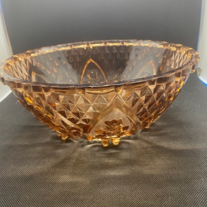 May include: A vintage amber glass bowl with a diamond pattern and a floral design on the bottom. The bowl has a scalloped edge and sits on three small feet.