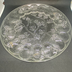 May include: A clear glass serving plate with a raised design of a pine tree with pine cones. The plate has a scalloped edge.