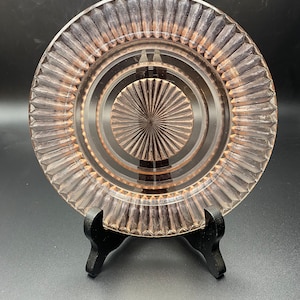 May include: A clear, pink-tinted glass plate with a ribbed outer edge and concentric circles in the center. The plate is displayed on a black stand against a dark background. The design features a sunburst pattern in the center.