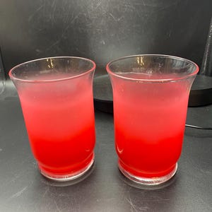 May include: Two clear glass candle holders with a red ombre effect. The candle holders are empty and sitting on a black surface.