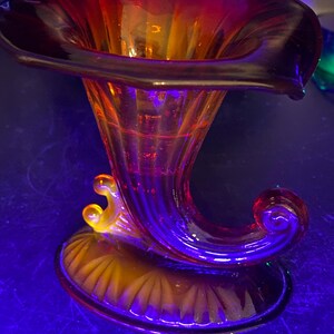 May include: A red and orange glass vase with a trumpet shape and a fluted base. The vase has a swirl design and a decorative handle.