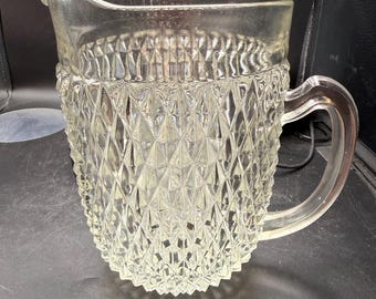 May include: A clear glass pitcher with a diamond pattern around the base and a curved handle. The pitcher has a spout for easy pouring. The glass is transparent, allowing the contents to be visible.