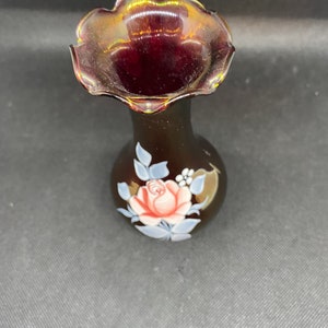 May include: A small, dark red glass vase with a ruffled top and a painted design of a pink rose with white and green leaves.