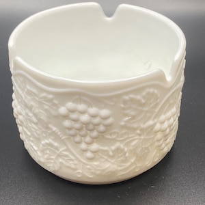 May include: White milk glass ashtray with a grapevine design. The ashtray has a raised rim and a wide, shallow bowl.