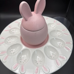 May include: A white ceramic Easter egg serving platter with bunny-shaped egg holders and a pink bunny-shaped egg holder in the center. The bunny has pink ears and a speckled finish. The platter has a pink base.