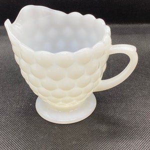 May include: White milk glass creamer with a textured, bubble-like design. The creamer has a handle and a round base.