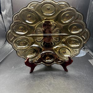 May include: A vintage amber-colored glass serving dish with a scalloped edge and starburst details. The dish is divided into sections and sits on a dark wooden stand. The dish is designed for serving appetizers or snacks.