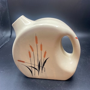 May include: A white ceramic pitcher with a brown and black design of cattails. The pitcher has a rounded body and a handle.