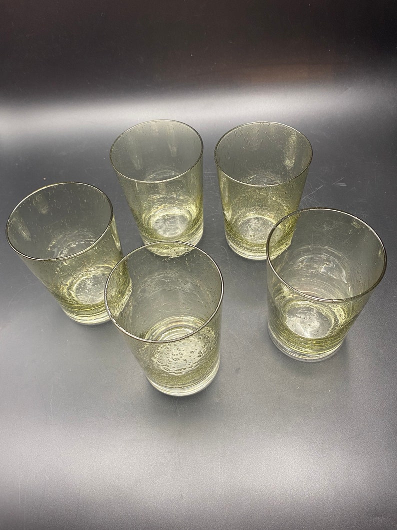 Puede incluir: Cinco vasos transparentes de vidrio con una superficie texturizada y burbujeante. Los vasos est&aacute;n vac&iacute;os y dispuestos en c&iacute;rculo sobre una superficie oscura.