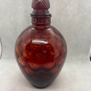 May include: A dark red glass bottle with a textured surface and a decorative stopper. The bottle has a rounded shape and a pointed top.