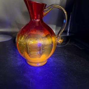 May include: A decorative glass pitcher with a vibrant color gradient. The top is a deep red, transitioning to a golden-yellow base. It features a curved handle and a spout for pouring.