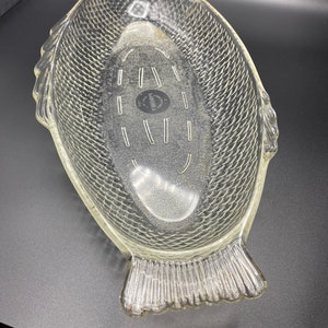 May include: A clear glass fish-shaped serving dish with a textured surface. The dish has a raised rim and a small circle in the center with the text "Lancaster Glass Corp. USA" printed on it.