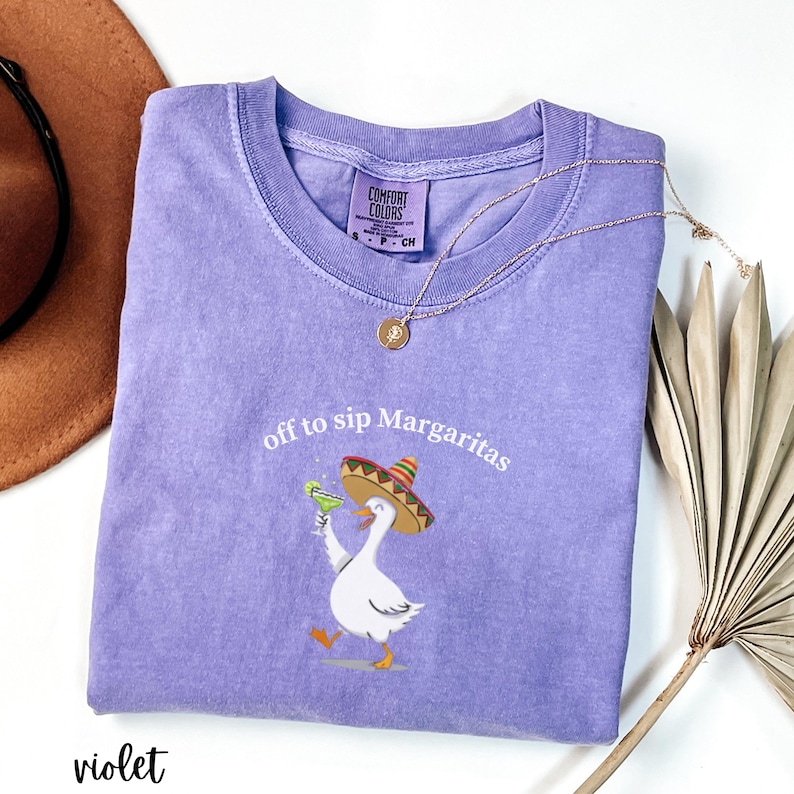 Peut inclure: T-shirt violet Comfort Colors avec l'inscription "off to sip Margaritas" et un dessin d'oie de dessin anim&eacute; portant un sombrero et tenant une margarita. Un collier dor&eacute; et un chapeau marron sont &eacute;galement visibles.