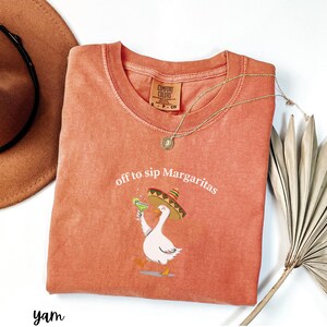 Peut inclure: Un t-shirt Comfort Colors de couleur corail avec l'inscription "off to sip Margaritas" et un dessin d'oie portant un sombrero et tenant une margarita. Un collier dor&eacute; et un chapeau marron sont &eacute;galement visibles.