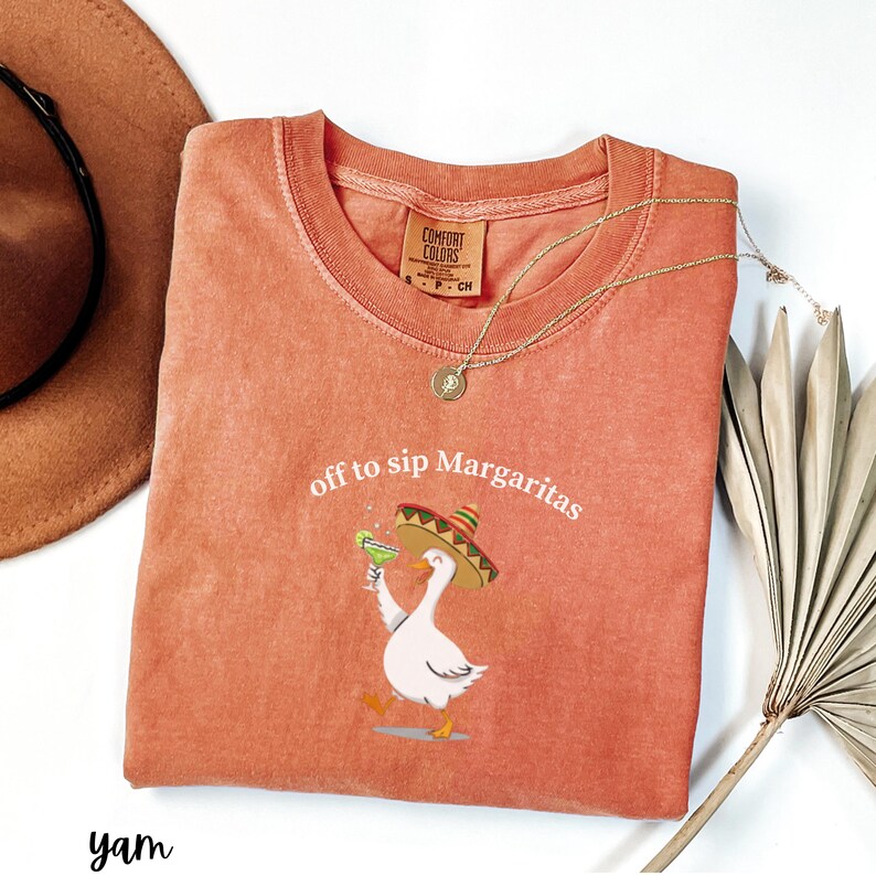 Peut inclure: Un t-shirt Comfort Colors de couleur corail avec l'inscription "off to sip Margaritas" et un dessin d'oie portant un sombrero et tenant une margarita. Un collier dor&eacute; et un chapeau marron sont &eacute;galement visibles.
