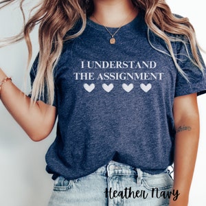 May include: A heather navy blue t-shirt with the text "I understand the assignment" and four white hearts.