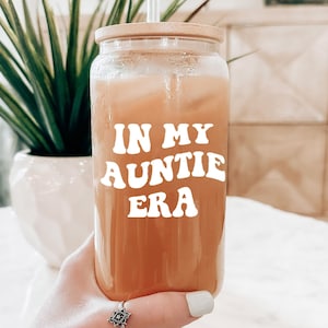 May include: A clear glass jar with a wooden lid and a straw. The jar is filled with a light brown liquid and has a white sticker with the text "IN MY AUNTIE ERA" in a retro font.