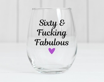 May include: A clear stemless wine glass with the words "Sixty & Fucking Fabulous" in black script. A small purple heart is below the text. The glass is designed for celebratory occasions and is suitable for wine or other beverages.