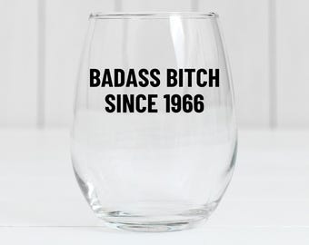 May include: A clear stemless wine glass with the text "BADASS BITCH SINCE 1966" in bold black letters. The glass is empty and sits on a white surface.