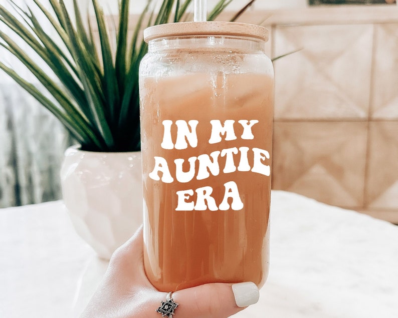May include: A clear glass jar with a wooden lid and a straw. The jar is filled with a light brown liquid and has a white sticker with the text "IN MY AUNTIE ERA" in a retro font.