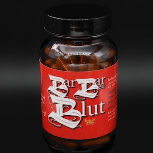 Könnte beinhalten: Eine klare Glasflasche mit schwarzem Deckel, gefüllt mit braunen Kapseln. Die Flasche hat ein rotes Etikett mit weißem Text, der "Bar Bar Blüt" lautet. Der Hintergrund ist schwarz.