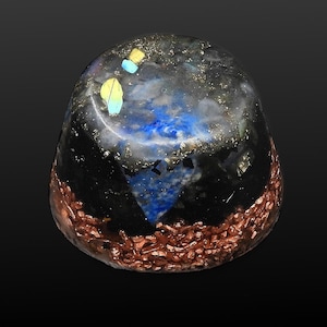 Op de afbeelding: Een eivormig orgonkristal met een koperen basis. Het kristal heeft blauwe, gele en turquoise accenten in een zwarte en heldere hars. De bovenkant heeft een gespikkelde uitstraling.