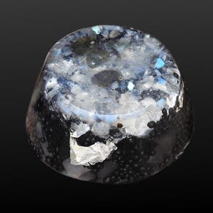 Könnte beinhalten: Ein dekoratives Objekt aus klarem Glas mit abgerundeter Form. Das Glas ist mit schwarzen, weißen und blauen Elementen gefüllt, wodurch ein strukturiertes Aussehen entsteht. Die Basis des Objekts ist dunkel und geht nach oben in hellere Farbtöne über.