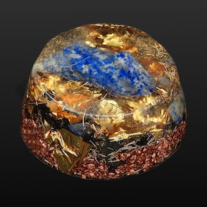 Può includere: Un fermacarte in resina a forma di cupola contenente lapislazzuli incastonati, scaglie di foglia d'oro e trucioli di rame. La base in resina trasparente rivela una composizione a strati di materiali blu, oro metallizzato e color rame.