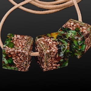 Puede incluir: Un collar único con tres colgantes cúbicos. Cada colgante es una mezcla de cobre y elementos verdes encapsulados en resina transparente. El collar está ensartado en un cordón marrón claro.