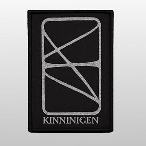 May include: A rectangular black patch featuring a silver embroidered design of a rounded rectangle intersected by diagonal lines. The word KINNINIGEN is embroidered in silver capital letters at the bottom of the patch, centred below the graphic.