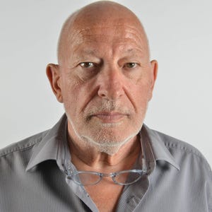 May include: A middle-aged person with a shaved head and light stubble, wearing a grey button-down shirt with a pointed collar. A pair of clear-framed glasses is hanging from the neckline of the shirt, resting against the chest area.