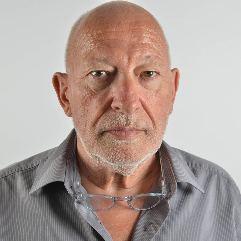 May include: A middle-aged person with a shaved head and light stubble, wearing a grey button-down shirt with a pointed collar. A pair of clear-framed glasses is hanging from the neckline of the shirt, resting against the chest area.