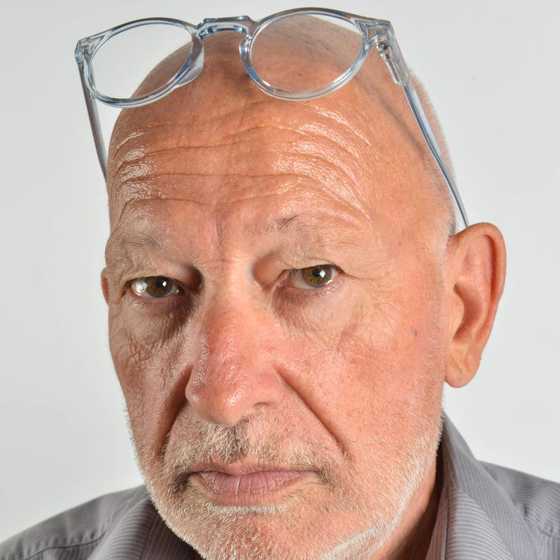 May include: A pair of clear, transparent plastic-framed eyeglasses resting on the forehead of a person with a shaved head, light skin, and a short, graying beard, wearing a light gray collared shirt with a subtle vertical pinstripe pattern.