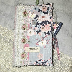 Pode incluir: Um diário feito à mão com um design floral. A capa apresenta uma mistura de padrões florais rosa, brancos e azuis, acabamentos em renda e detalhes de pérolas. A palavra "JOURNAL" está impressa na capa.