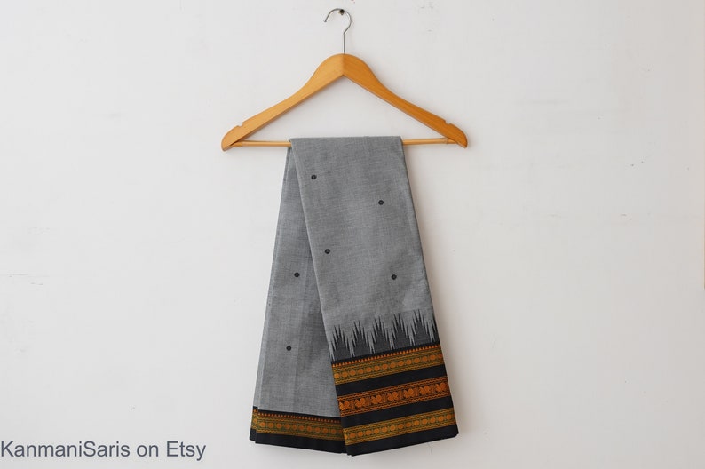 May include: A gray cotton saree with black polka dots and a black and gold border. The saree is hanging on a wooden hanger against a white background.