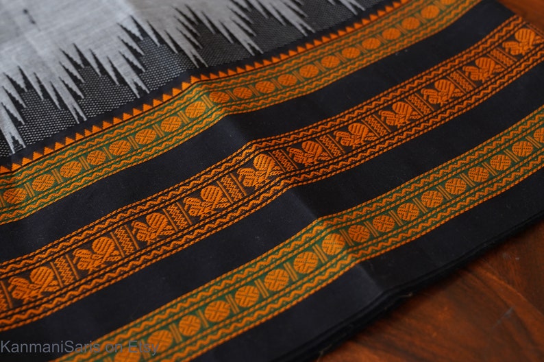 May include: Close-up of a black fabric with orange and green woven stripes. The fabric has a repeating pattern of circles and stylized flowers.