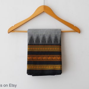 May include: A black and grey cotton saree with a gold and green border. The saree is hanging on a wooden hanger against a white background.