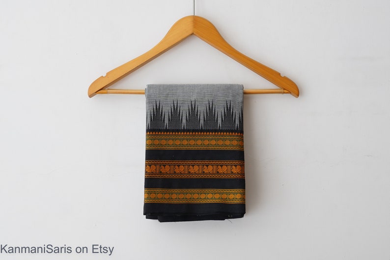 May include: A black and grey cotton saree with a gold and green border. The saree is hanging on a wooden hanger against a white background.