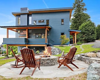 Può includere: Vista esterna di una moderna casa a due piani con rivestimento blu e un ponte di legno. La casa ha un braciere con sedie Adirondack disposte intorno. La scena è ambientata in una giornata di sole con un cielo azzurro.