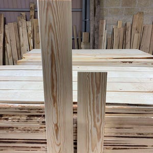 May include: Two rectangular wooden planks with visible wood grain. The planks are light brown and stand upright against a backdrop of stacked timber. The wood appears to be freshly cut and ready for use in a woodworking project.