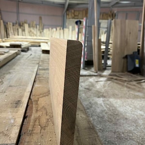 May include: A close-up of a light brown, unfinished wooden board with a rounded edge. The board is thick and has visible wood grain. The image is taken in a workshop setting, with other wood pieces in the background.