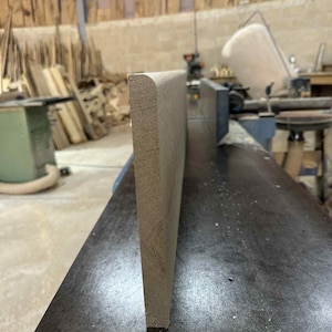 May include: A light brown wooden plank with a rounded edge, seen in close-up. The plank stands upright on a dark metal surface, possibly in a workshop. The wood grain is visible. The overall aesthetic is rustic and natural.